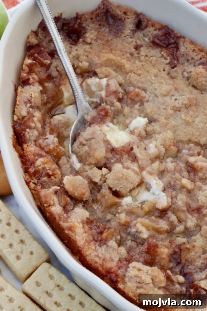 Warm baked apple pie dip in an oven-safe casserole dish, ready to be served with a spoon. The golden-brown crumble topping and bubbling apple filling are visible.