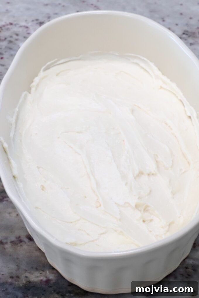 Smooth cream cheese filling spread evenly in the bottom of a glass casserole dish, forming the base layer of the dip.