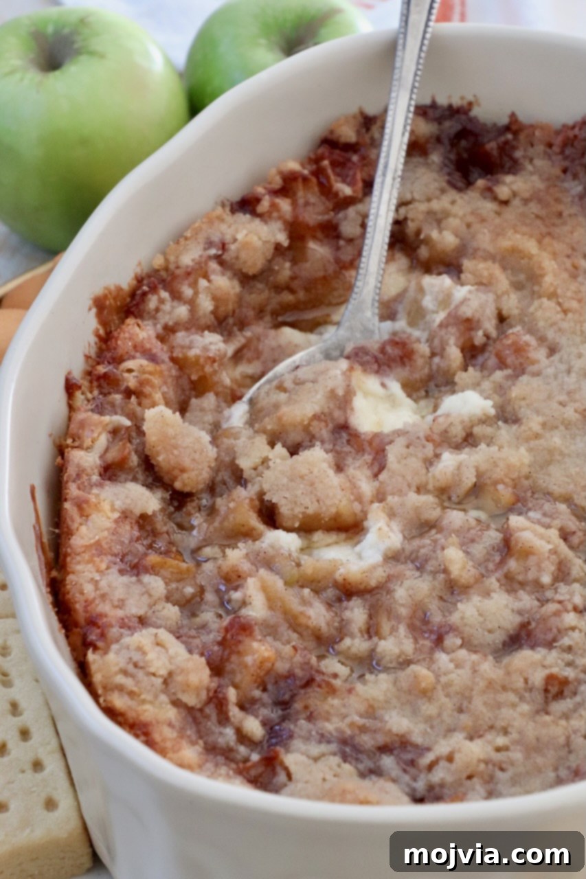 Close-up of baked apple pie dip in a casserole dish, showing the warm, spiced apple filling and the golden, crumbly topping with a serving spoon.