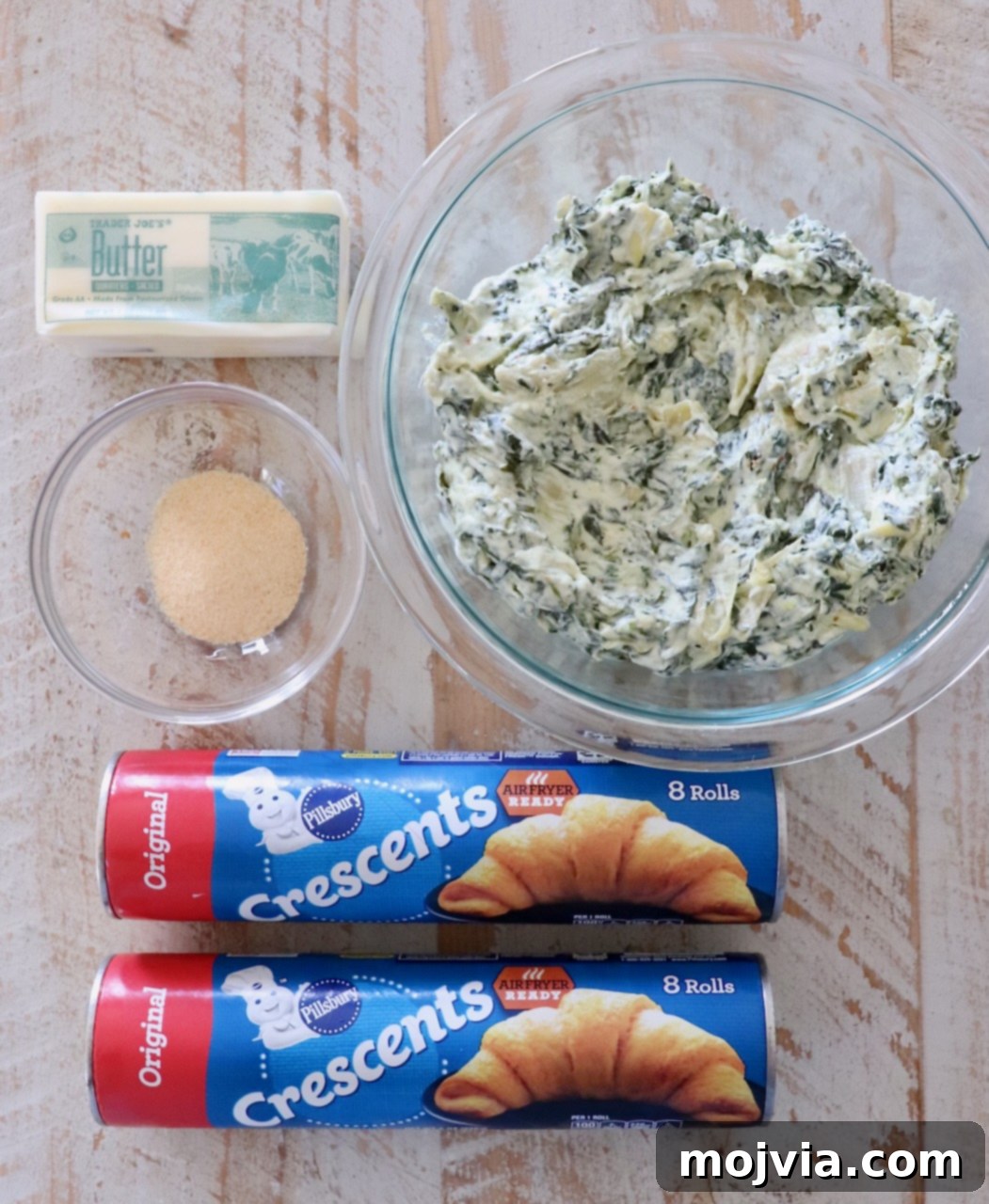 Savory Spinach Swirls 3 Ingredients for spinach pinwheels arranged on a white wooden board, including crescent roll dough, spinach artichoke dip, butter, and garlic powder