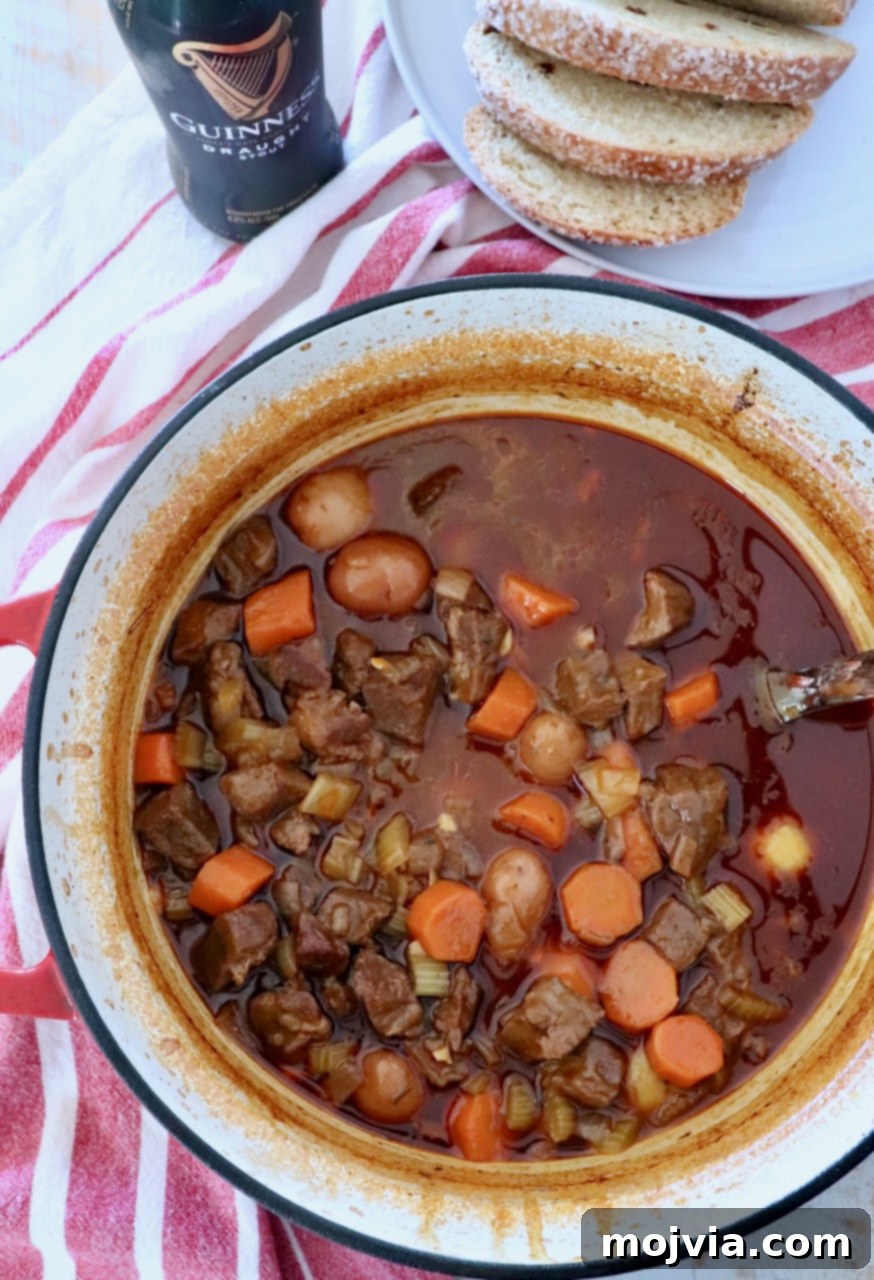 Beef stew simmering in a large cast iron pot next to a bottle of Guinness and sliced bread