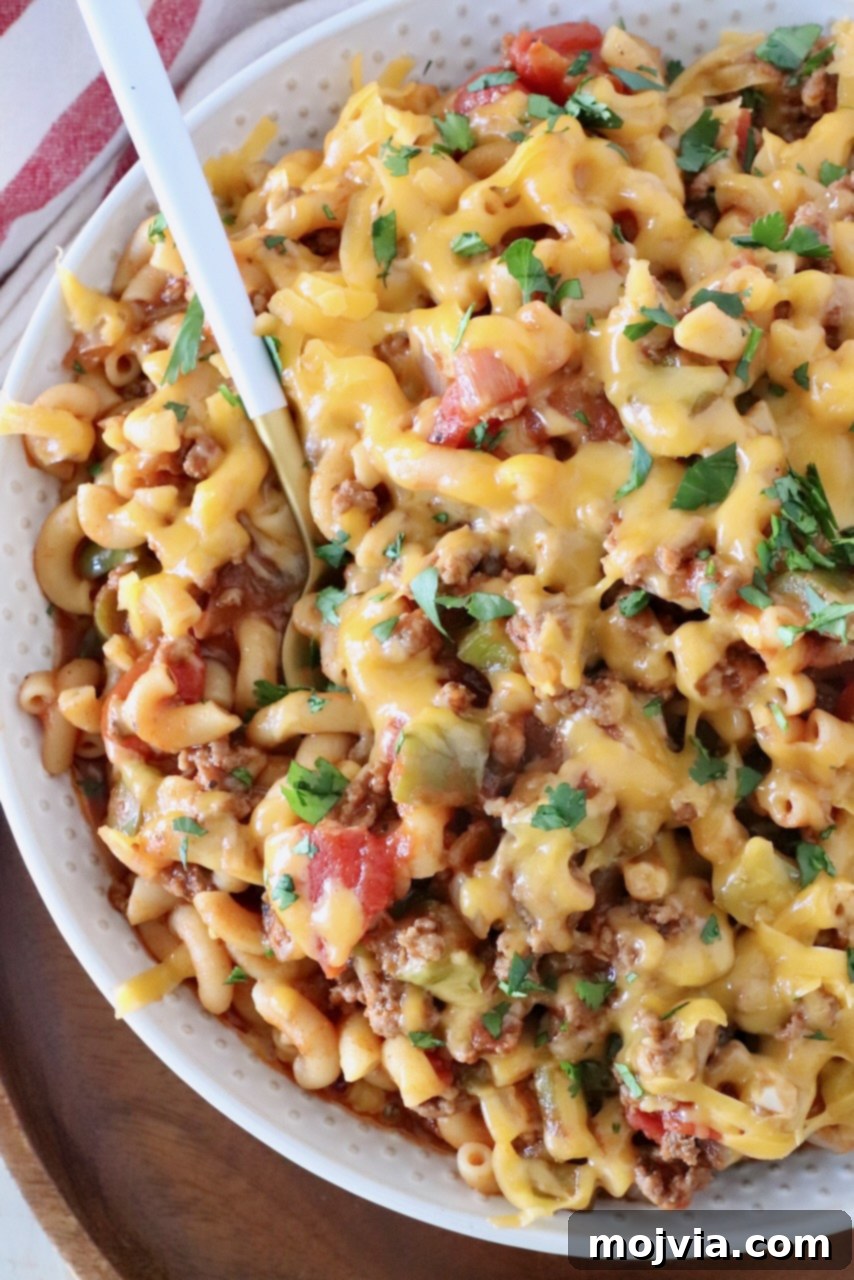 A close-up of prepared American Goulash in a bowl, generously covered with melted shredded cheddar cheese and a sprig of fresh parsley, emphasizing its rich and appealing texture.