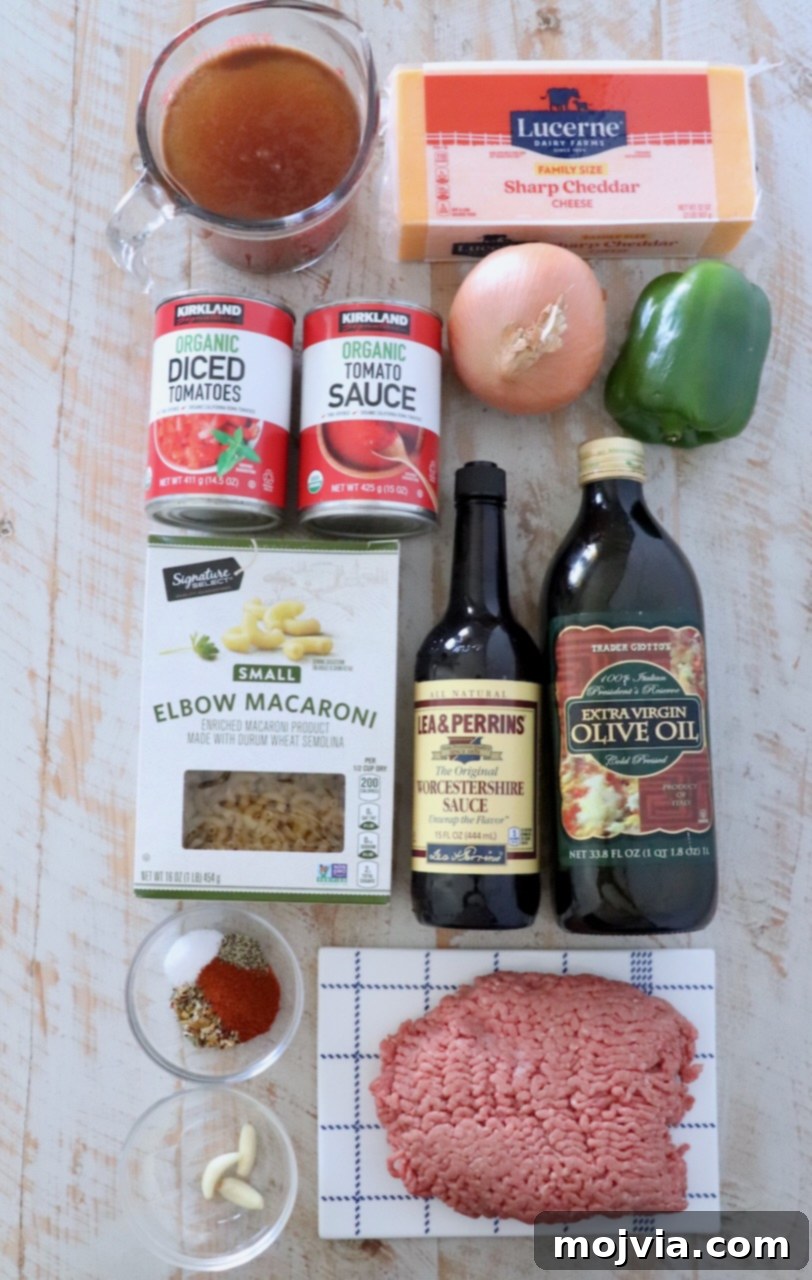 A collection of fresh ingredients for American goulash laid out on a rustic white wooden board, including ground beef, diced tomatoes, pasta, bell peppers, onions, and various spices.