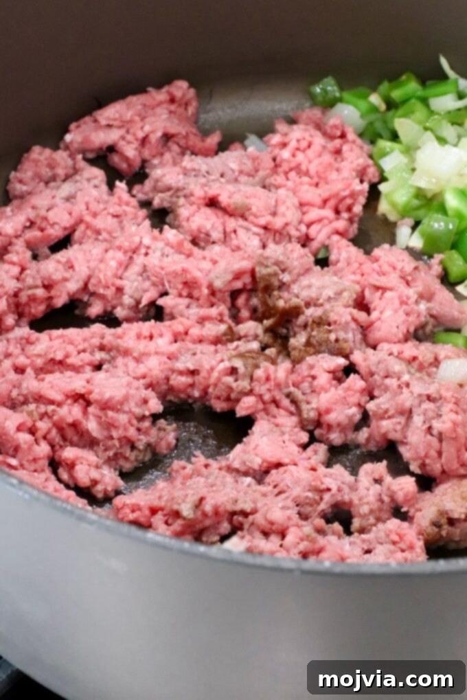 Ground beef browning in a large pot alongside softened diced onions and bell peppers, ready to be seasoned.