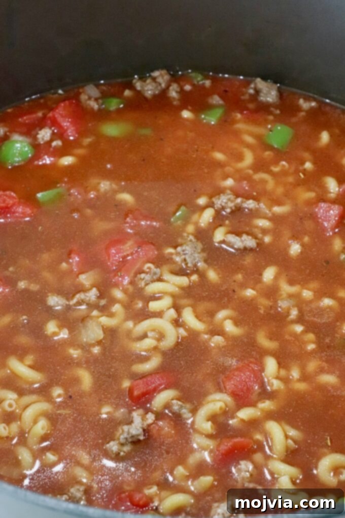Elbow macaroni, ground beef, and vegetables submerged in beef broth and tomato sauce, simmering in a large pot.