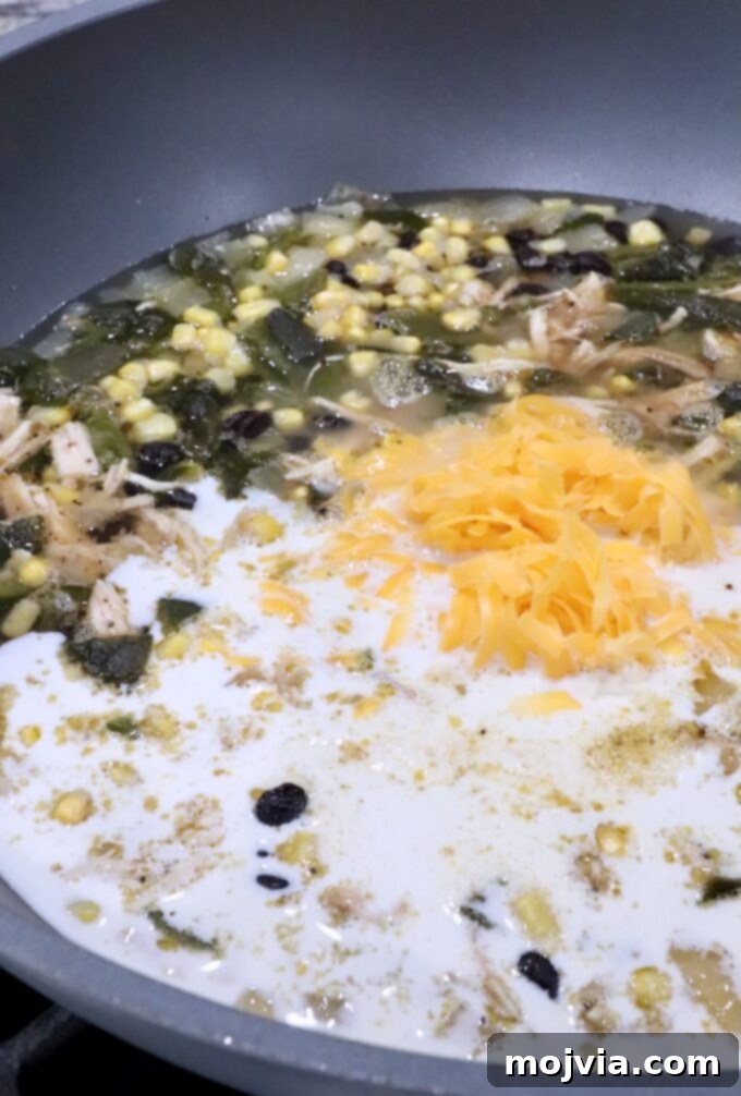 Roasted Poblano Chicken Soup 10 Shredded cheddar cheese and heavy cream being added to the simmering chicken poblano soup in a large pot.