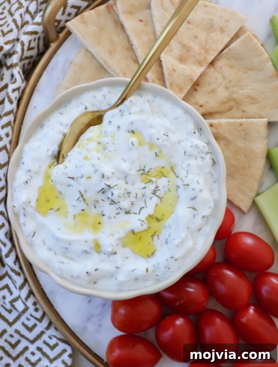 Creamy Greek Cucumber Garlic Dip 2 Tzatziki sauce in a bowl with a spoon, drizzled with olive oil, accompanied by slices of pita bread and cherry tomatoes on the side, highlighting its freshness and appeal.