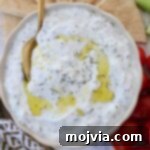 Homemade Tzatziki Sauce in a bowl, garnished with a spoon, olive oil drizzle, and surrounded by fresh pita bread slices and cherry tomatoes for serving.