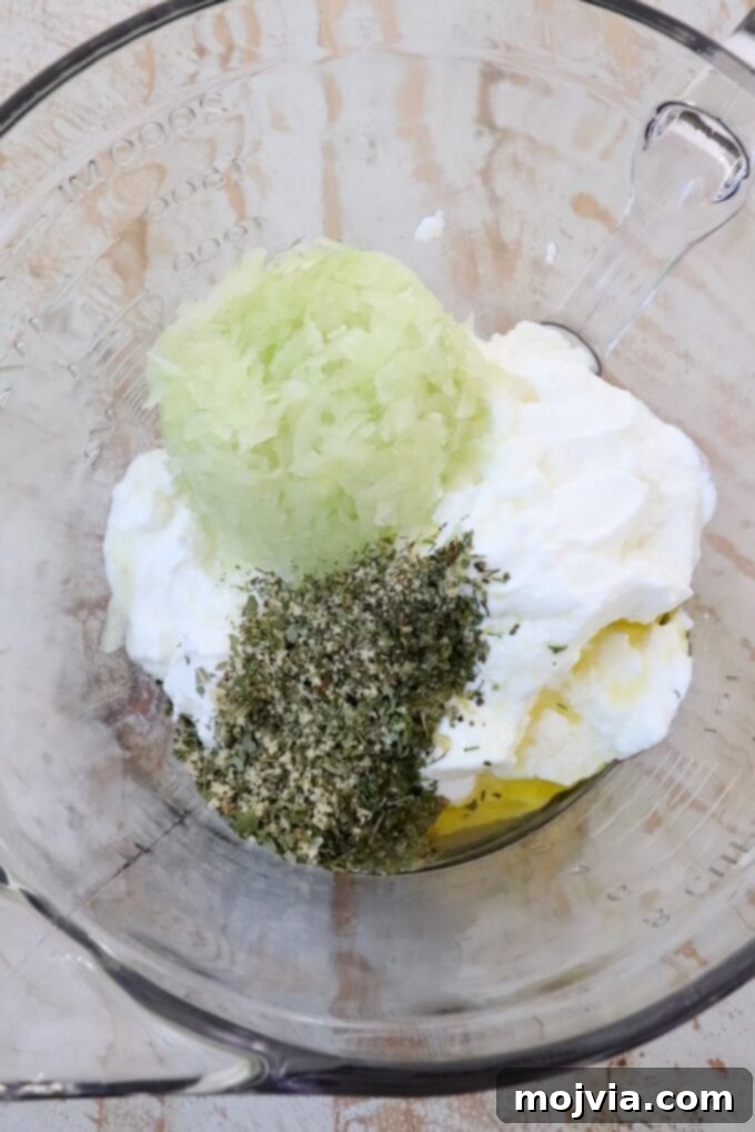 Creamy Greek Cucumber Garlic Dip 4 A large glass bowl filled with the base ingredients for tzatziki sauce: creamy Greek yogurt, finely grated cucumber, olive oil, and a vibrant blend of dried Greek herbs and spices.