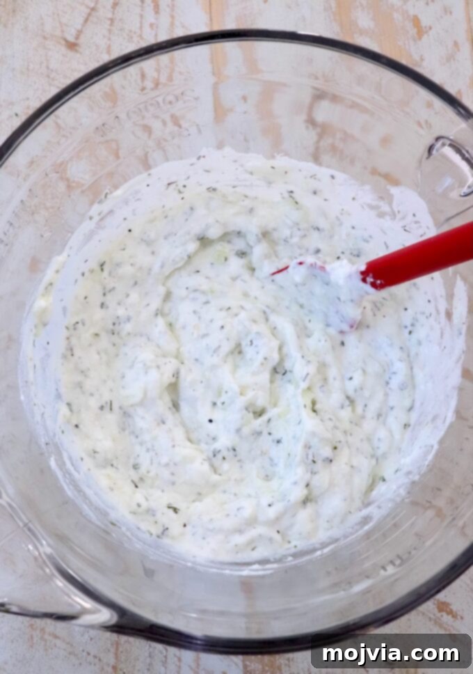 Creamy Greek Cucumber Garlic Dip 5 Freshly mixed tzatziki sauce in a large glass bowl, being stirred with a red spatula, showcasing its creamy and well-combined texture.