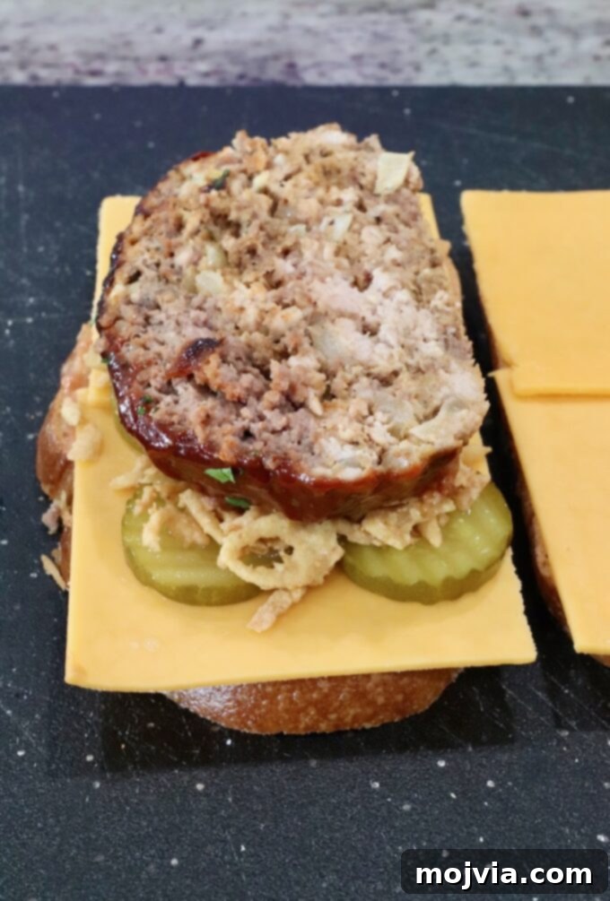 slice of meatloaf on top of cheese, pickles, and fried onion strings, on a piece of bread