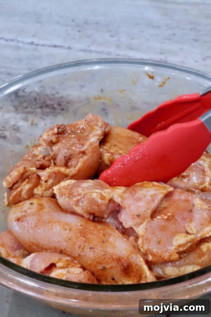 Sizzling Air Fryer Chicken Fajitas 4 Boneless chicken thighs being tossed with fajita seasoning, olive oil, and lime juice in a glass bowl using red kitchen tongs.