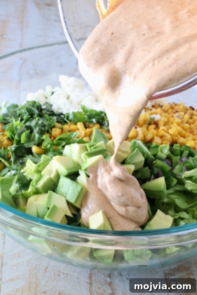 Esquites Style Corn Salad 8 corn, avocado, cotija cheese, cilantro and chopped lettuce in a large glass bowl with a creamy dressing poured over it out of a glass measuring cup