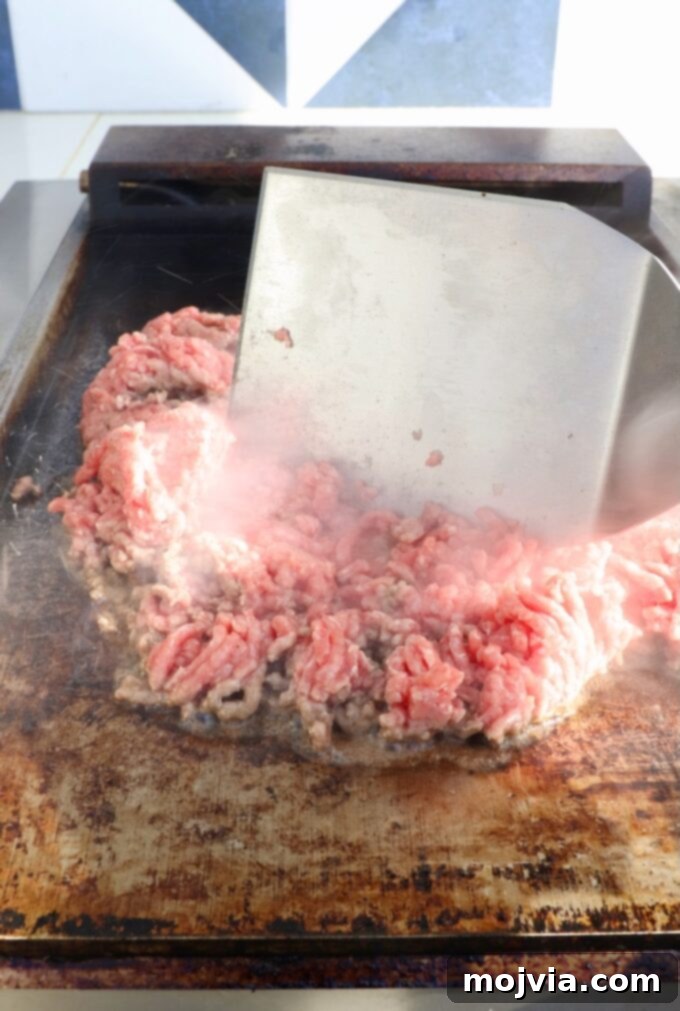 The Ultimate Chopped Cheese 4 Ground beef cooking on a flat top griddle, being chopped with a large metal spatula