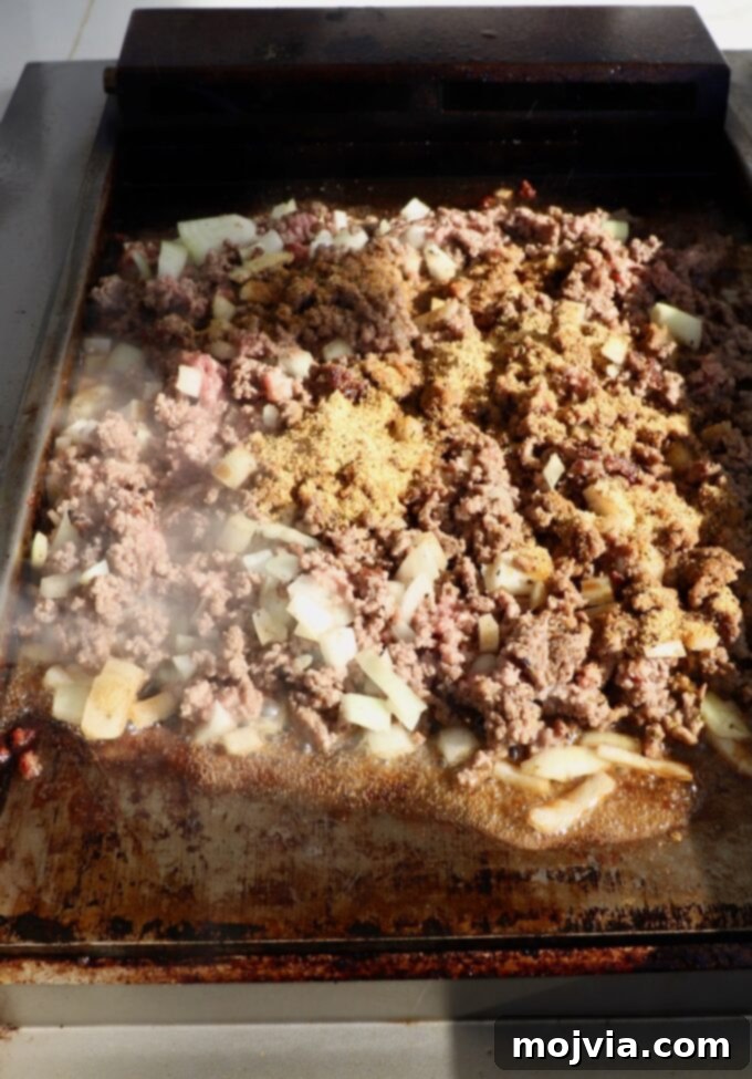 The Ultimate Chopped Cheese 5 Seasoned ground beef and diced onions being chopped together on a flat top griddle