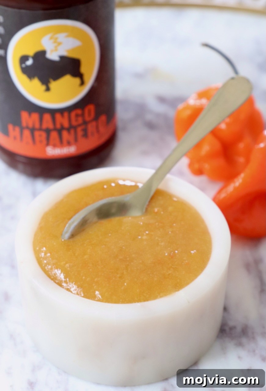 Sweet and spicy homemade mango habanero sauce in a bowl with a spoon, flanked by fresh habaneros and a bottle of store-bought sauce