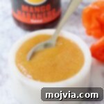 A small bowl of mango habanero sauce with a spoon, accompanied by fresh habanero peppers and a bottle of sauce in the background