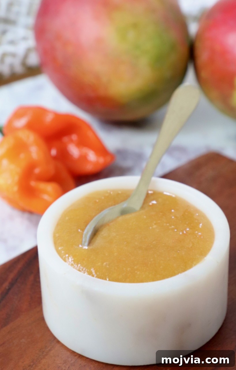 A serving of mango habanero sauce in a small bowl, with a spoon and fresh mangoes and habaneros in the background