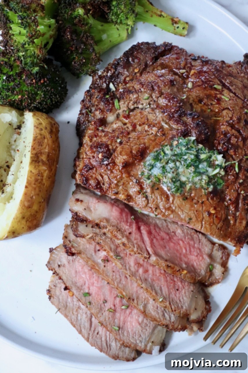 Air Fryer Steakhouse Steak 2 partially sliced ribeye steak on a plate with herb butter on top, cooked broccoli and a baked potato on the side