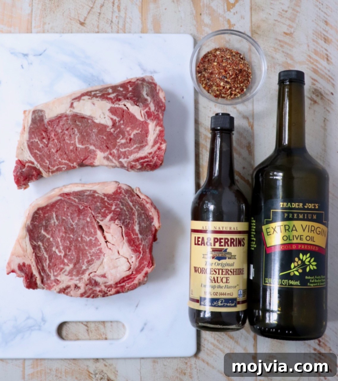 Air Fryer Steakhouse Steak 3 raw ribeye steaks on a cutting board, next to a bottle of Worcestershire sauce, a bottle of olive oil, and steak seasoning in a small glass bowl