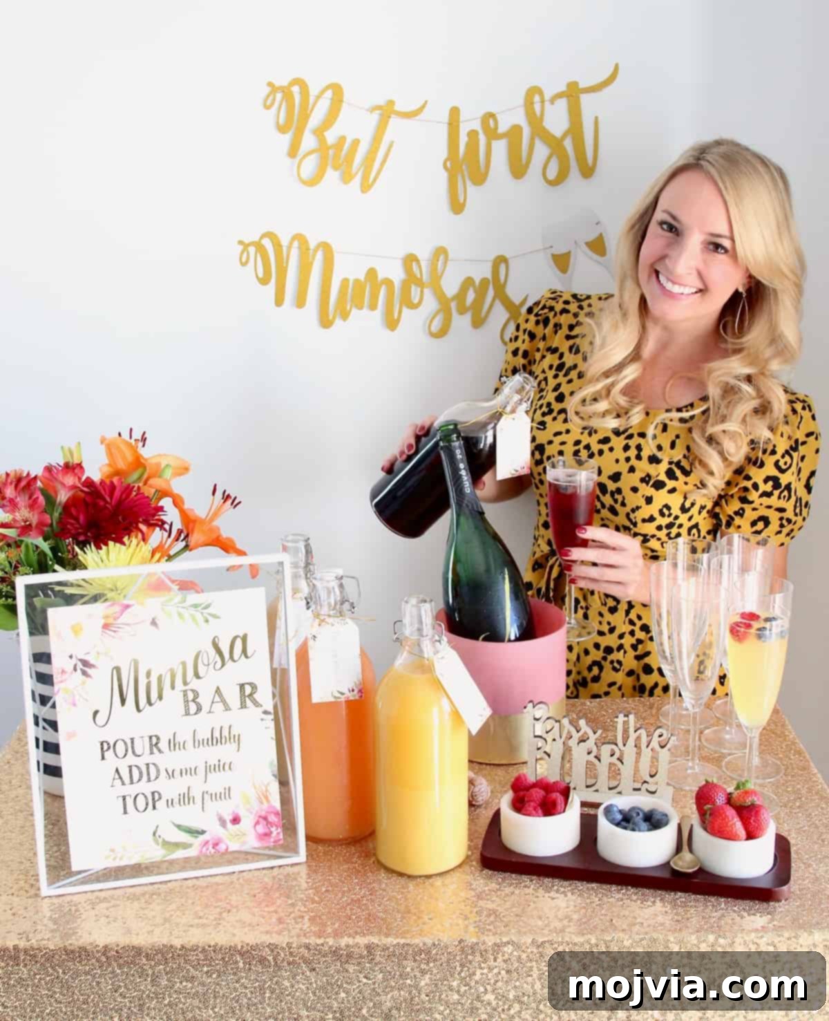 A woman elegantly pouring juice into a champagne flute, standing behind a beautifully arranged mimosa bar, ready for guests to customize their drinks.