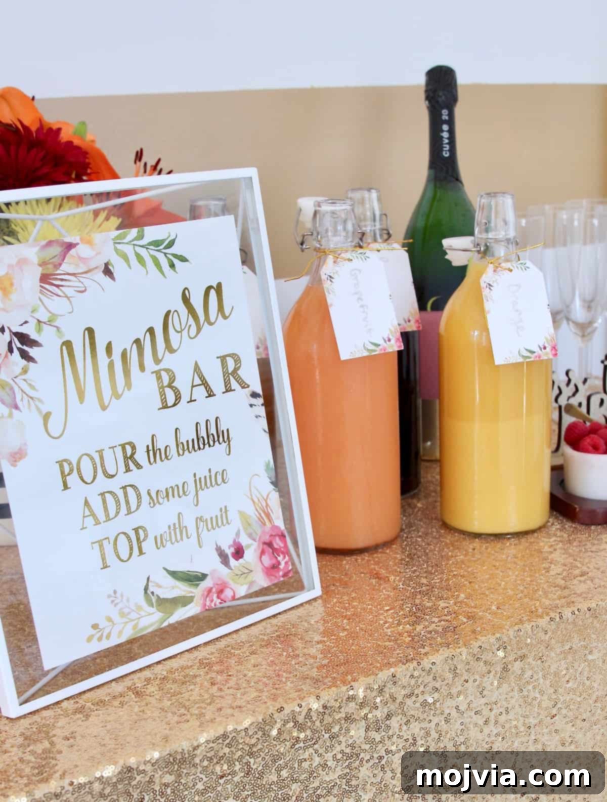 A festive mimosa bar setup featuring a gold sequin tablecloth, an elegant 'But First Mimosas' sign, and an array of juices in clear glass bottles.