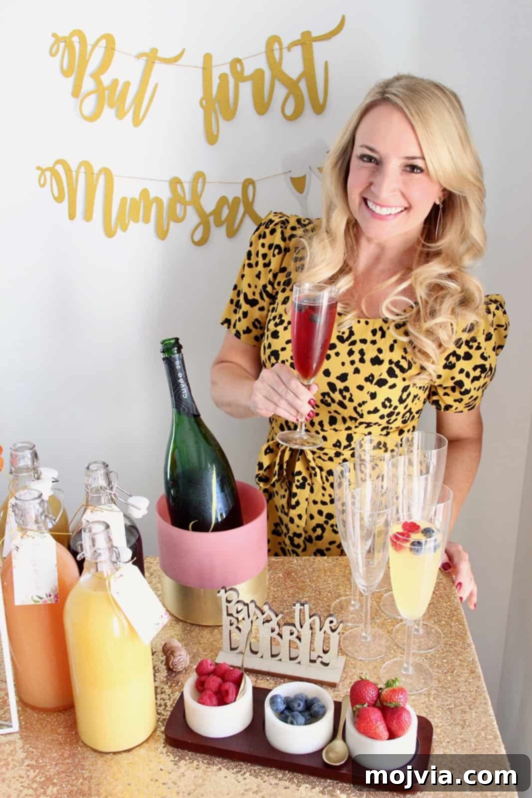 A woman smiling as she holds a freshly made mimosa cocktail, standing proudly behind her well-appointed mimosa bar at a party.