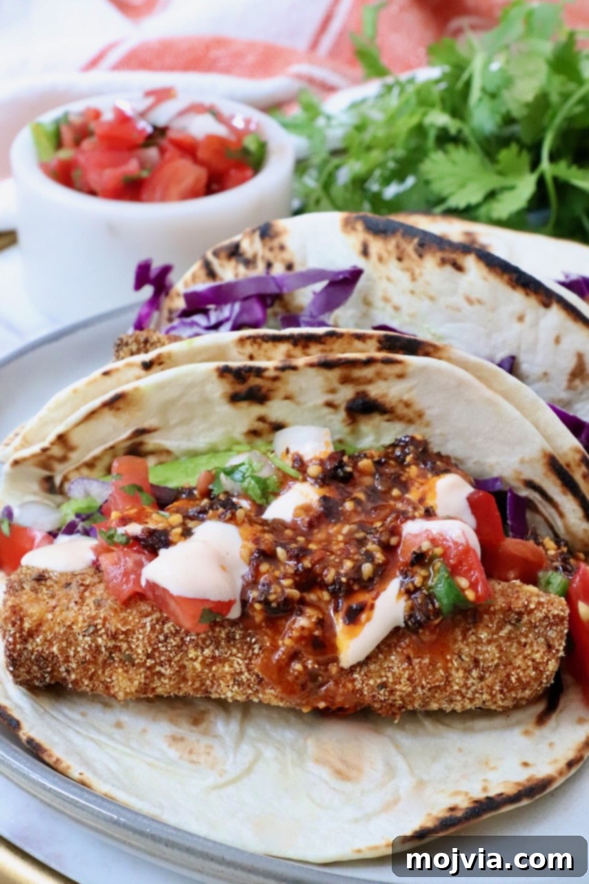A close-up of a perfectly constructed fish taco, featuring crispy breaded fish, vibrant red cabbage, fresh cilantro, a dollop of creamy sauce, and diced tomatoes, all nestled in a warm tortilla. A small dish of salsa and more fresh cilantro are blurred in the background, enhancing the fresh appeal.
