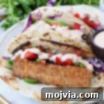 Three tacos with crispy fish, purple cabbage, diced tomatoes, and creamy sauce on a tortilla. With fresh cilantro in the background