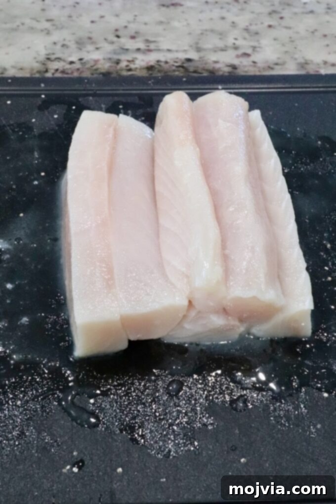 Raw mahi mahi fish fillets, expertly sliced into uniform strips, rest on a sleek black cutting board, ready for the next stage of preparation. The pristine white fish contrasts beautifully with the dark surface.