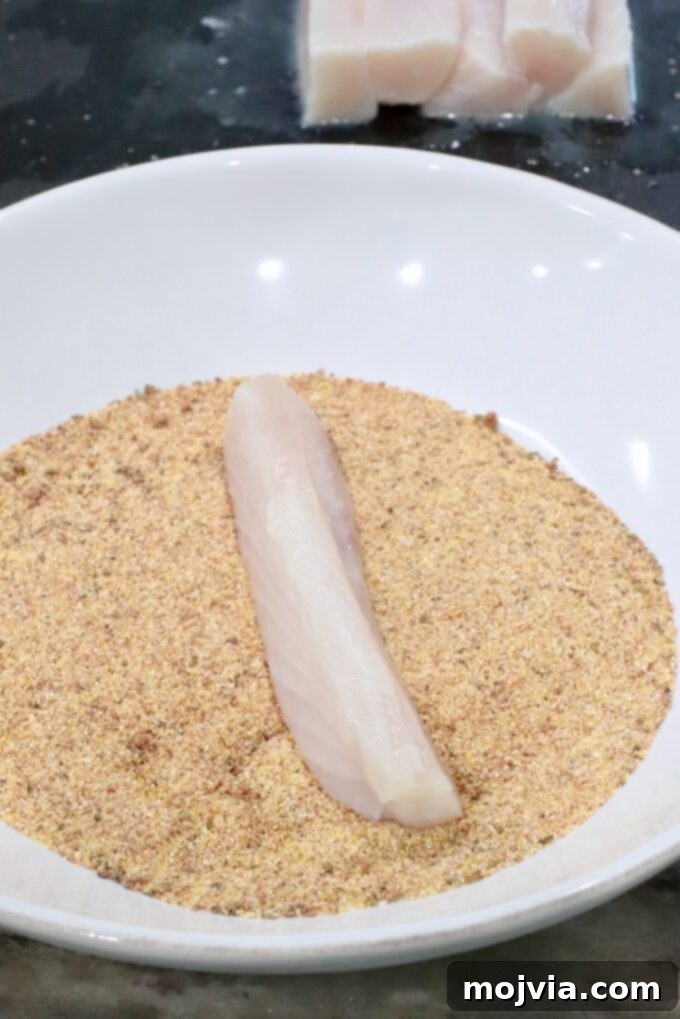 A pristine raw fish fillet is gently placed on a bed of seasoned cornmeal within a clean white bowl, poised for thorough breading. In the background, additional fish fillets await their turn on a dark, contrasting surface, ready for the next step in the recipe.