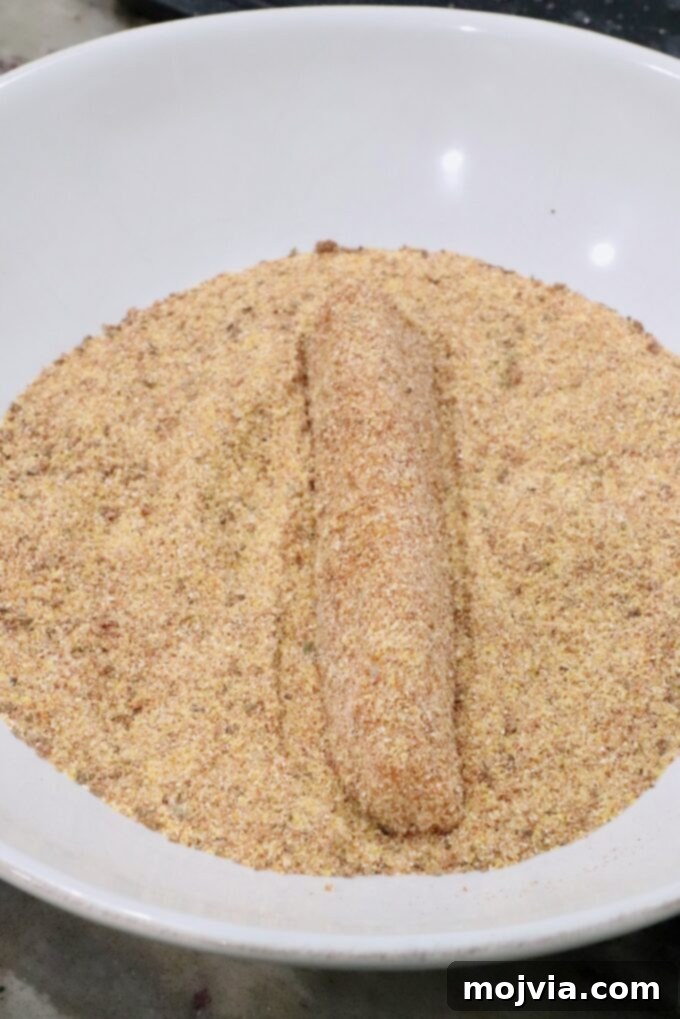 A perfectly breaded strip of fish is meticulously arranged on a bed of seasoned cornmeal within a pristine white bowl. The golden coating promises a crispy texture, indicating it's ready for the air fryer.