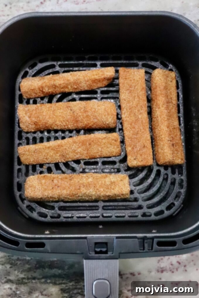 Six golden-brown, breaded fish strips are neatly arranged in a single layer inside an air fryer basket, positioned on a clean kitchen counter. Each piece is spaced perfectly for optimal air circulation, promising an even and crispy cook.