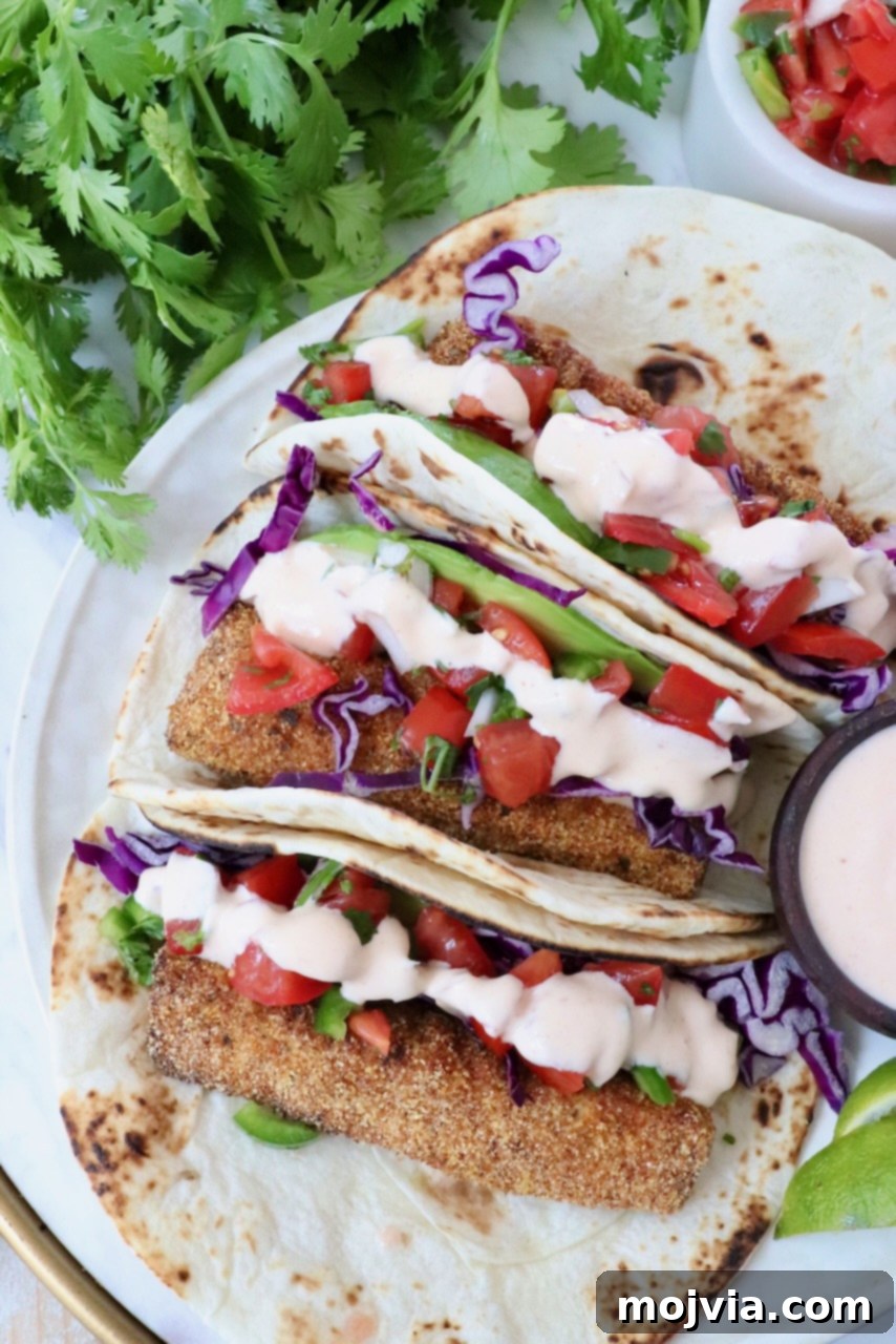 A vibrant and appetizing close-up of three prepared fish tacos, artfully arranged. Each taco features a golden, crispy fish fillet, a generous topping of vivid purple cabbage, freshly diced tomatoes from pico de gallo, and a creamy, zesty sauce, all nestled within a soft tortilla. The tacos are elegantly garnished with sprigs of fresh cilantro, emphasizing their fresh and flavorful appeal.