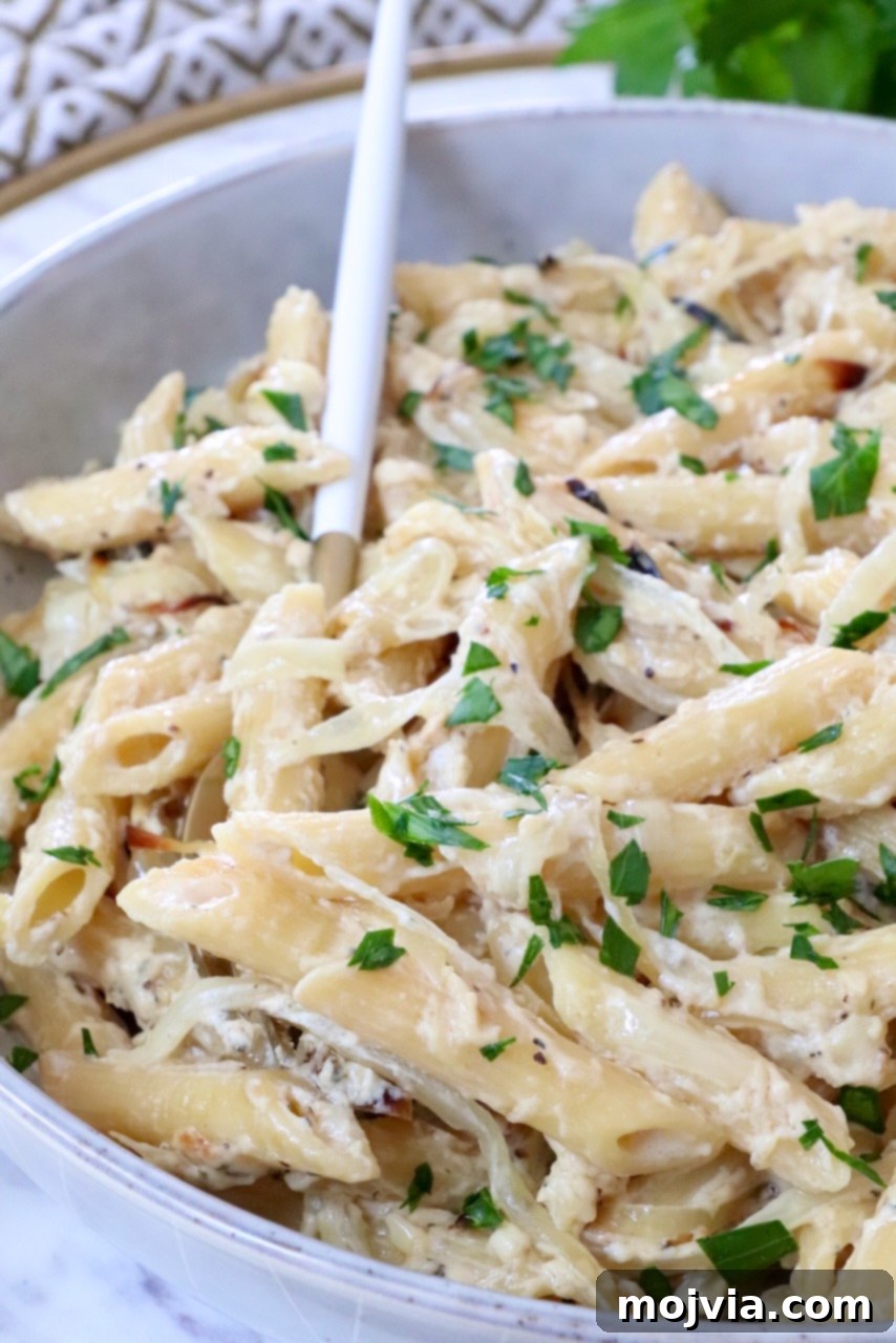 Golden French Onion Boursin Pasta 2 Creamy penne pasta topped with fresh parsley in a white bowl, accompanied by a white and gold spoon.