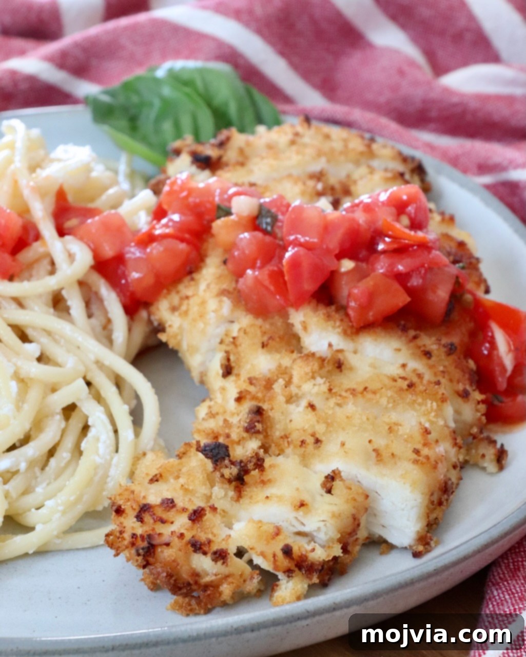 Sliced piece of crispy garlic chicken on a bed of creamy pasta, generously topped with vibrant tomato bruschetta, garnished with fresh basil.