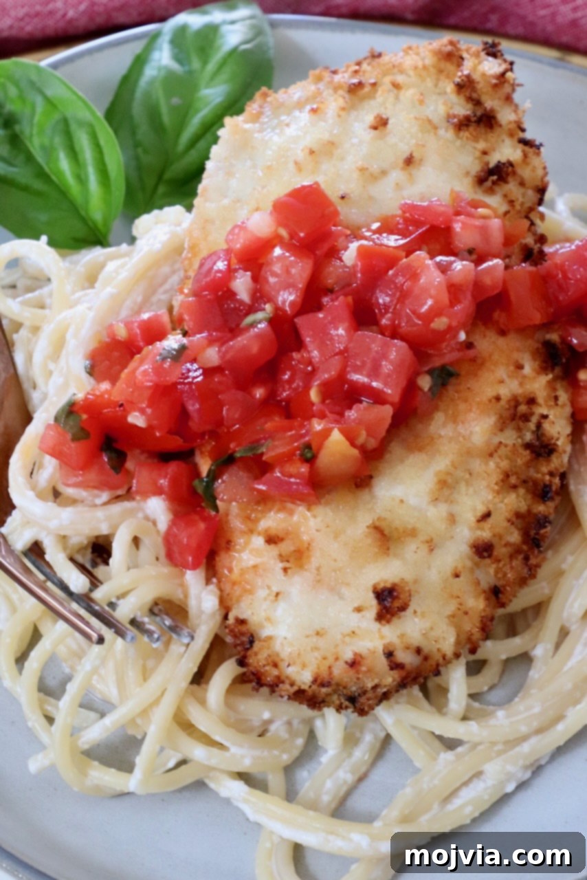 A beautifully plated dish featuring a golden-brown crispy chicken cutlet resting on a bed of creamy spaghetti, generously topped with fresh tomato bruschetta, with vibrant basil leaves scattered in the background.