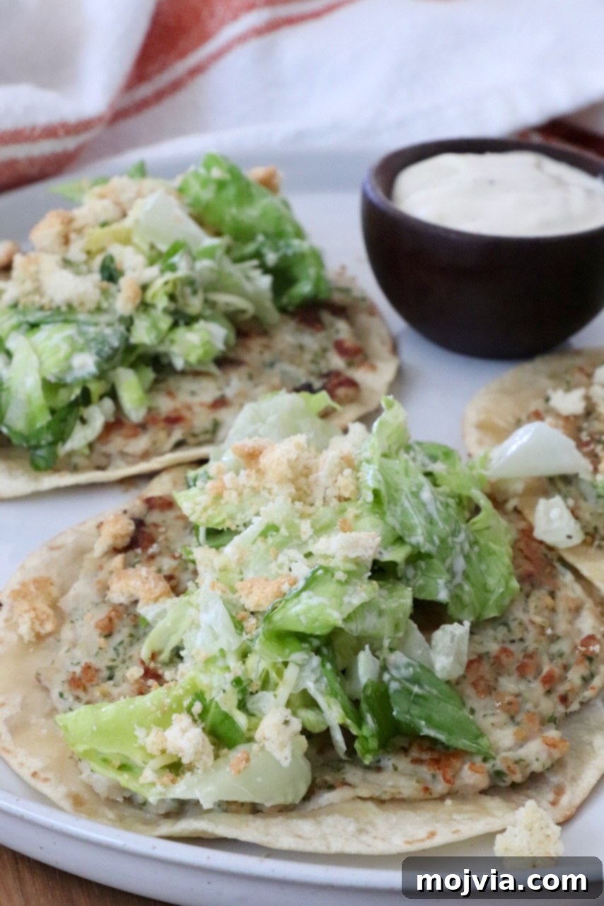 Two smash chicken tacos topped with leafy green salad and crumbled croutons on a white plate. A small bowl of creamy dipping sauce is on the side.
