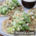 Two smash chicken tacos topped with leafy green salad and crumbled croutons on a white plate. A small bowl of creamy dipping sauce is on the side.