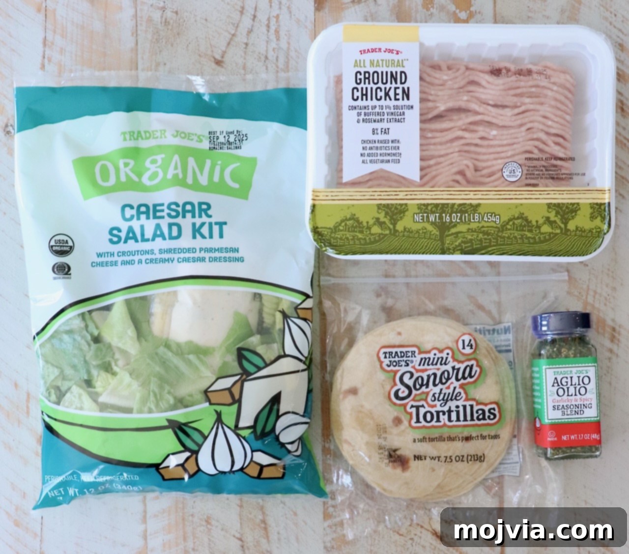 Trader Joe’s products on a wooden surface: organic Caesar salad kit, ground chicken, mini Sonora tortillas, and Aglio Olio seasoning blend.