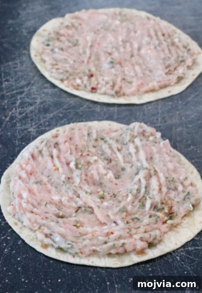 Two tortillas on a dark cutting board, each topped with a spread of raw ground chicken mixed with herbs and spices