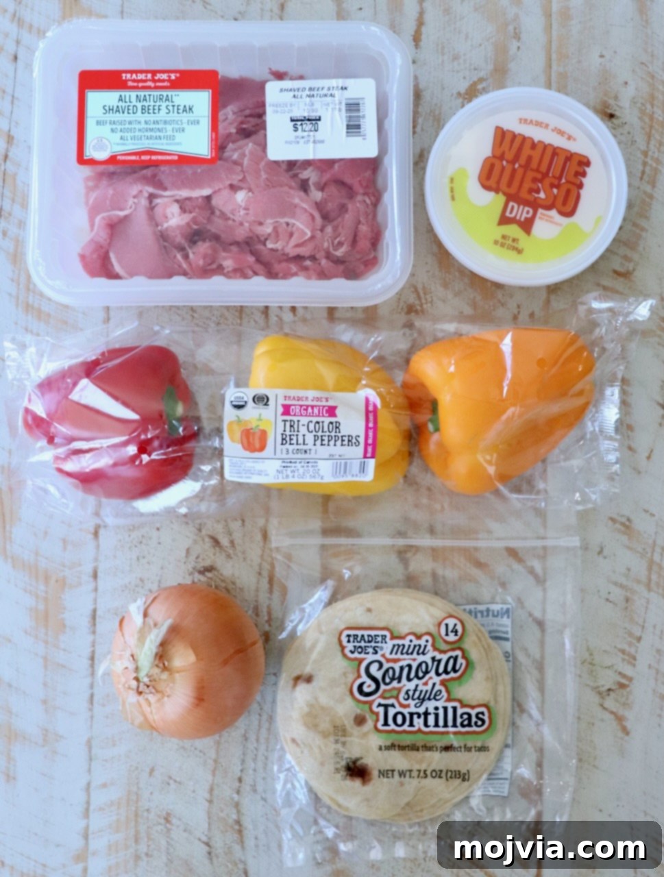 Assorted Trader Joe's ingredients on a wooden surface: shaved beef steak, white queso dip, tri-color bell peppers, a yellow onion, and mini tortillas.