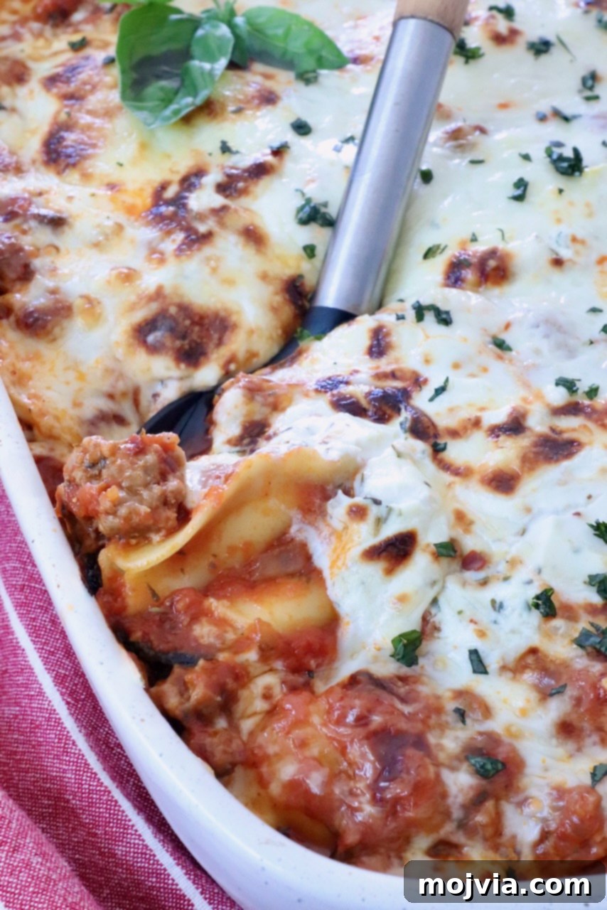 Cheesy Baked Ravioli Delight 2 Close-up of a baked ravioli casserole in a white dish, topped with melted cheese and herbs. A serving spatula lifts a portion, showing layers of pasta and meat sauce.
