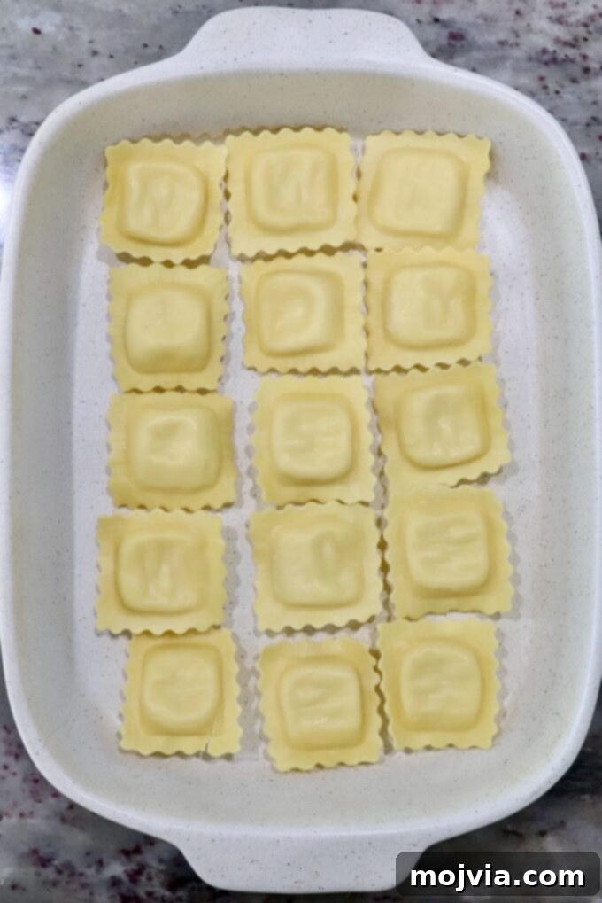 Cheesy Baked Ravioli Delight 7 White baking dish with 15 uncooked ravioli in neat rows on a marble countertop.