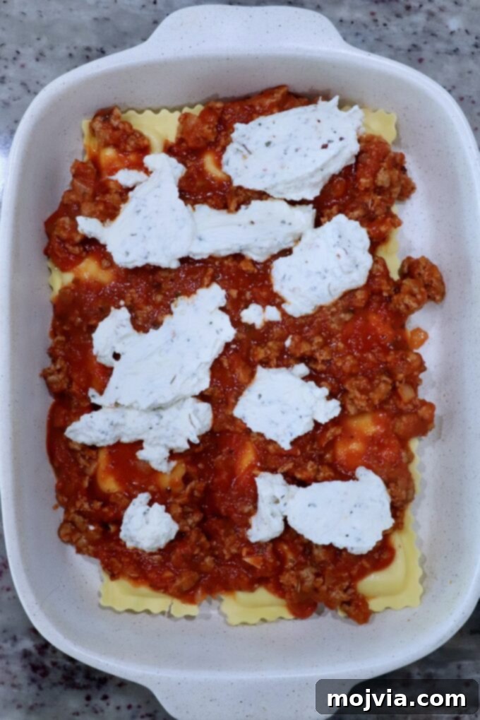 Cheesy Baked Ravioli Delight 8 Baked ravioli preparation in a white dish on a speckled countertop. Layers of pasta are topped with meat sauce and dollops of ricotta cheese.