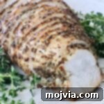 Sliced herb-crusted cooked turkey breast on a white plate, surrounded by fresh green herbs.