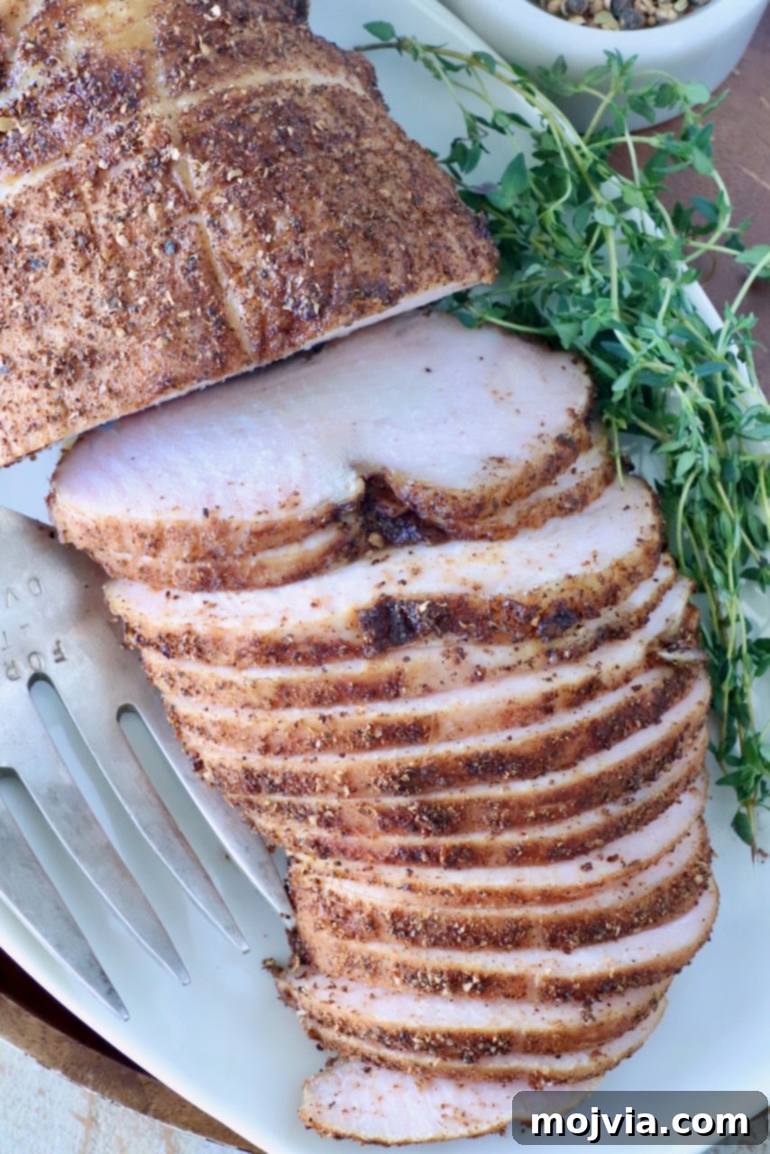 Sliced smoked turkey breast on a white platter, seasoned with spices. Fresh herbs are placed beside it, along with a serving fork.