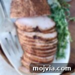 Sliced smoked turkey breast on a white platter, seasoned with spices. Fresh herbs are placed beside it, along with a serving fork.
