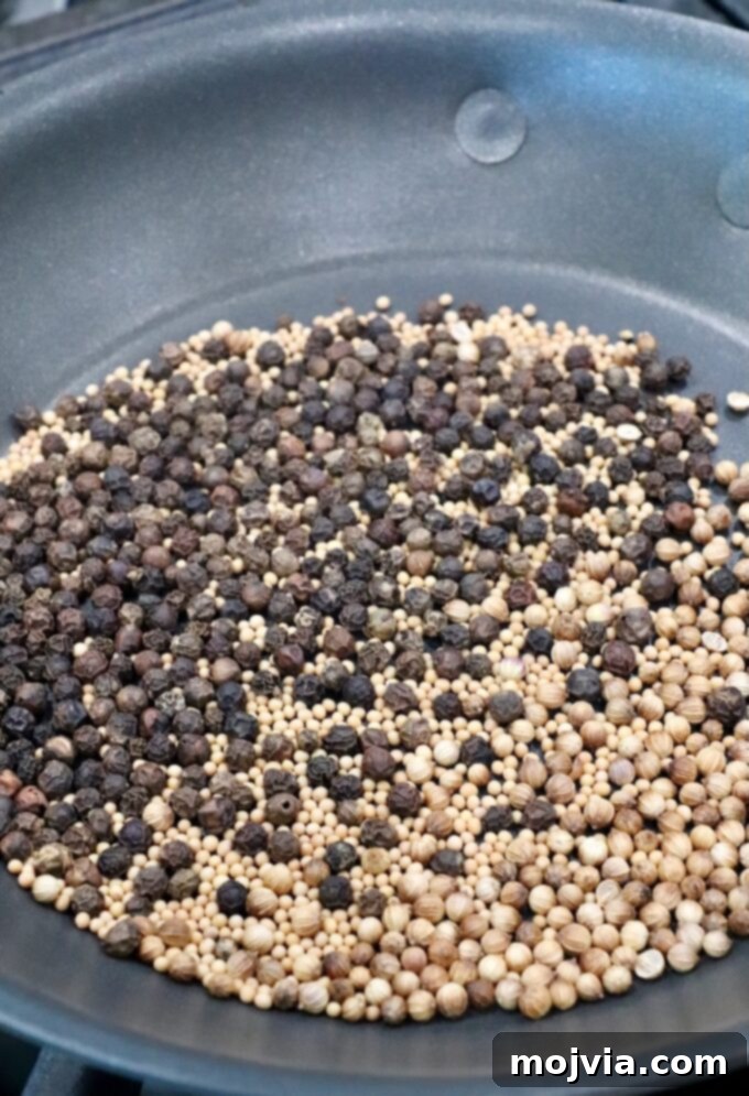 Assorted whole spices, including black peppercorns, mustard seeds, and coriander seeds, are being dry roasted in a dark frying pan.