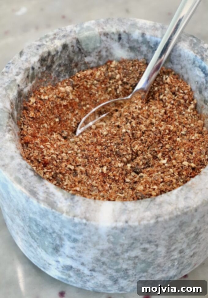 A close-up of a marble mortar filled with a coarse, mixed spice blend. A shiny metal spoon is partially submerged in the spices